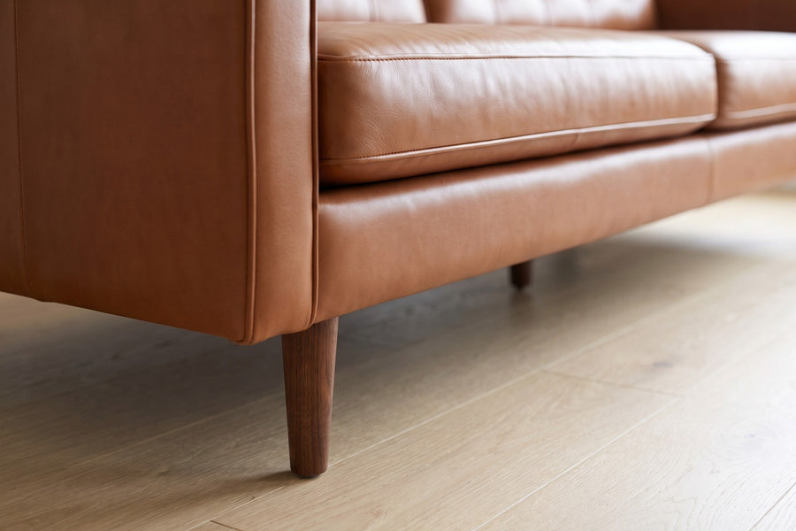 Amboise Cognac Button-Tufted Leather Sofa: close-up of a round tapered walnut-finish leg under the front corner of the sofa, on a pale oak floor