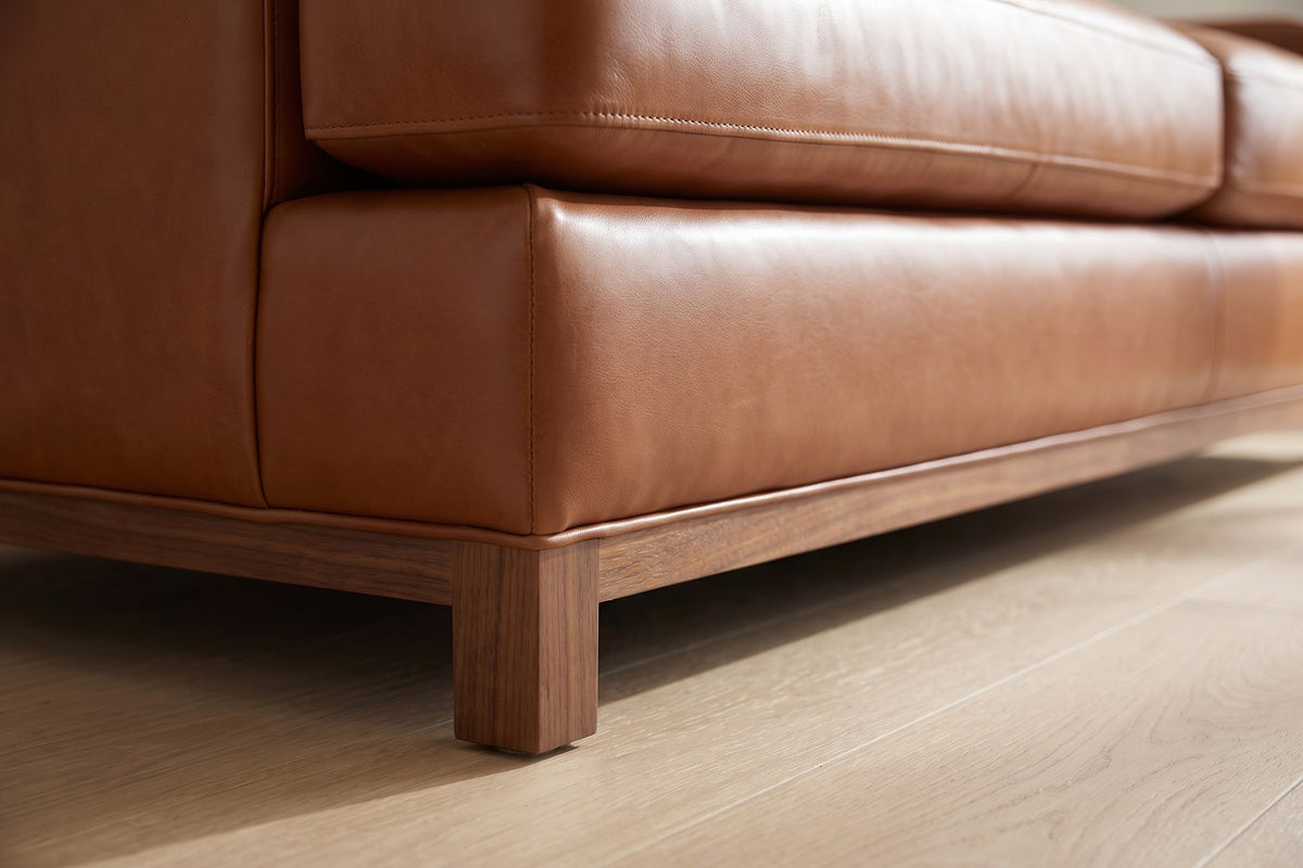 Arcachon Cognac Deep Leather Sofa: close-up of the slim walnut-finish base rail and a short square leg under the front corner, on a pale oak floor