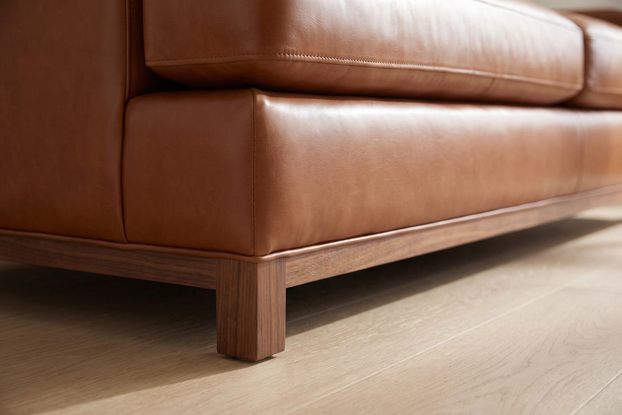 Arcachon Cognac Deep Leather Sofa: close-up of the slim walnut-finish base rail and a short square leg under the front corner, on a pale oak floor