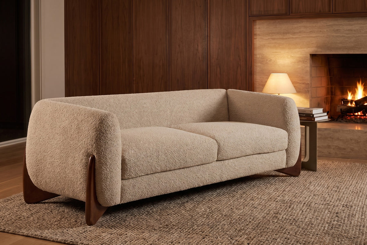 Evening. Beside a lit fire and a table lamp, the almond bouclé looks warm on a woven rug.