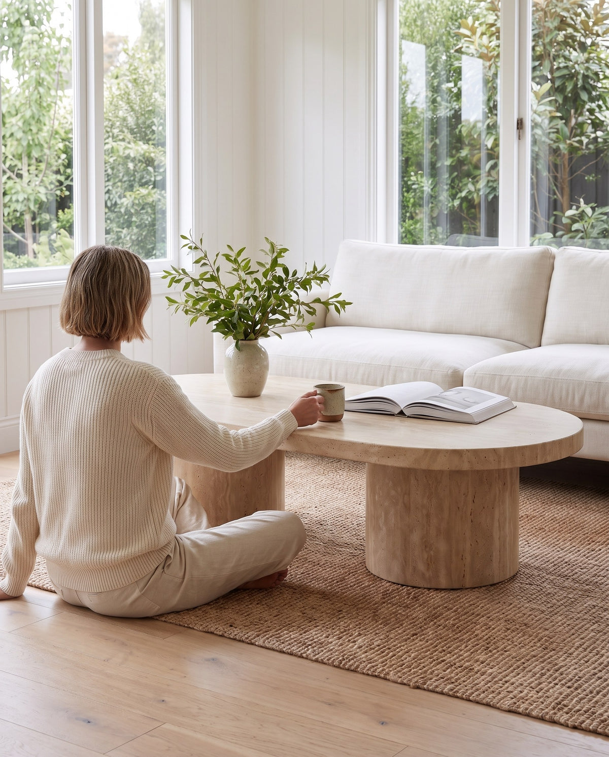 A person sitting on the rug at the Bandol travertine coffee table with a cup and an open book (concept image)