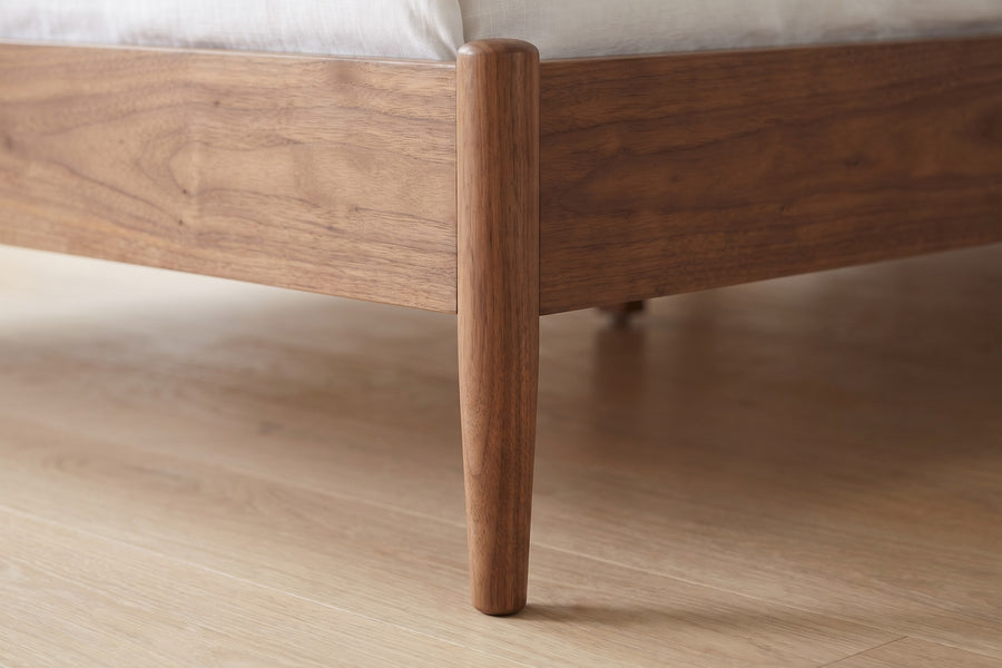 Bergerac Walnut and Cane Bed: close-up of a slim tapered walnut leg under the foot corner of the side rail, on a pale oak floor