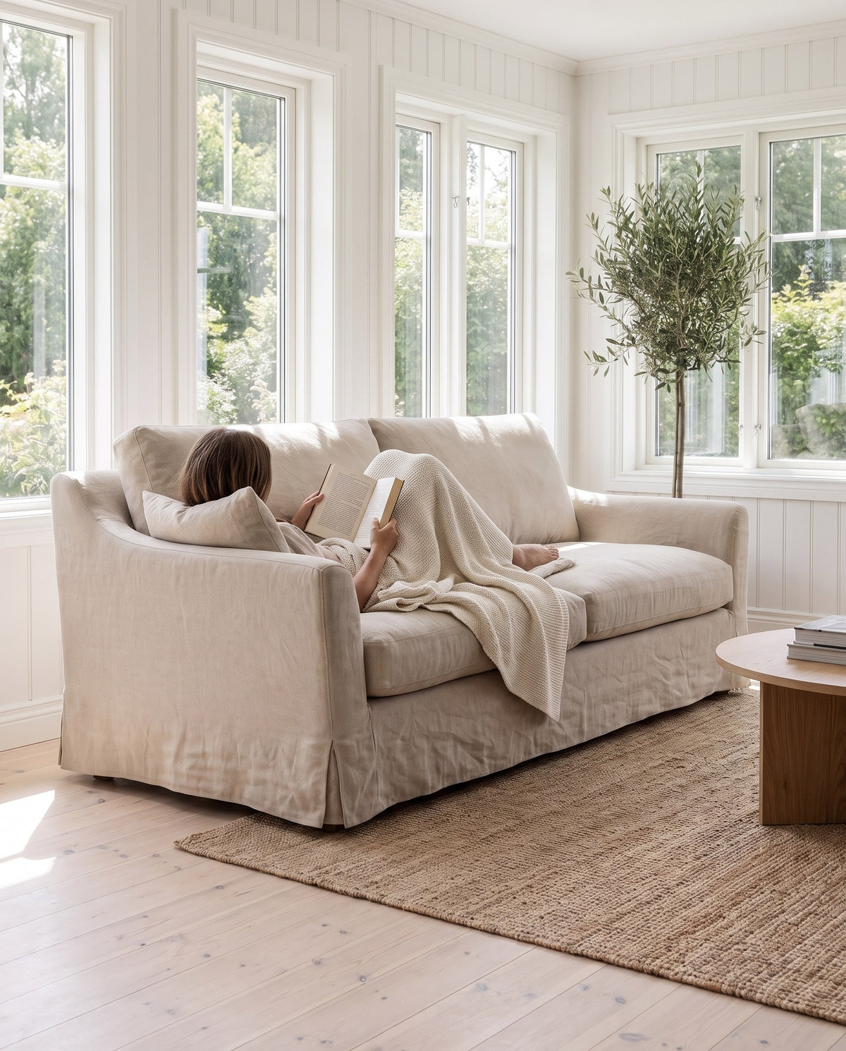 A person curled up reading in the corner of the Germain natural linen slipcovered sofa (concept image)