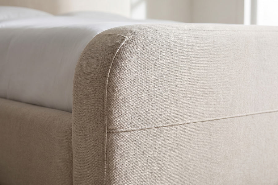 Giverny Oatmeal Upholstered Low Bed: close-up of a rounded top corner of the headboard where it flows down into the side rail, showing the oatmeal woven texture and the seam