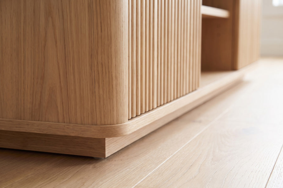 Gordes Fluted Oak Media Console: close-up of a softly rounded front corner of the case and the recessed plinth base just above a pale oak floor