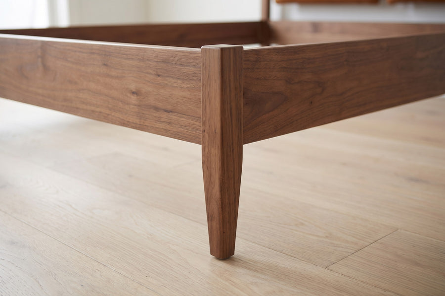 Honfleur Walnut Bed with Cognac Leather Cushions: close-up of the corner where the walnut side rail meets the low footboard and a slim tapered leg, on a pale oak floor