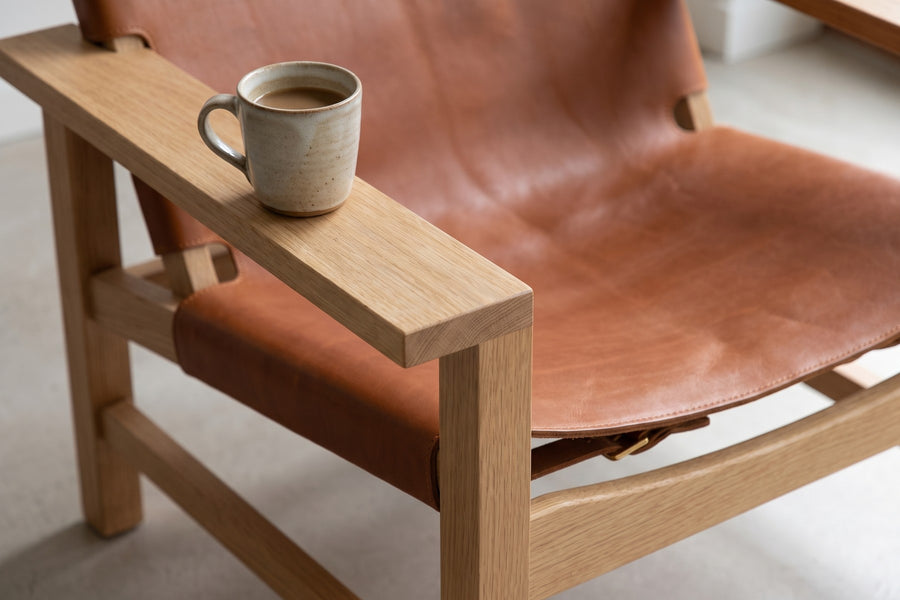 Lourmarin Cognac Leather Sling Chair on Natural Oak: close-up of a wide flat natural oak arm with a ceramic cup of coffee resting on it, the cognac seat sling beside it