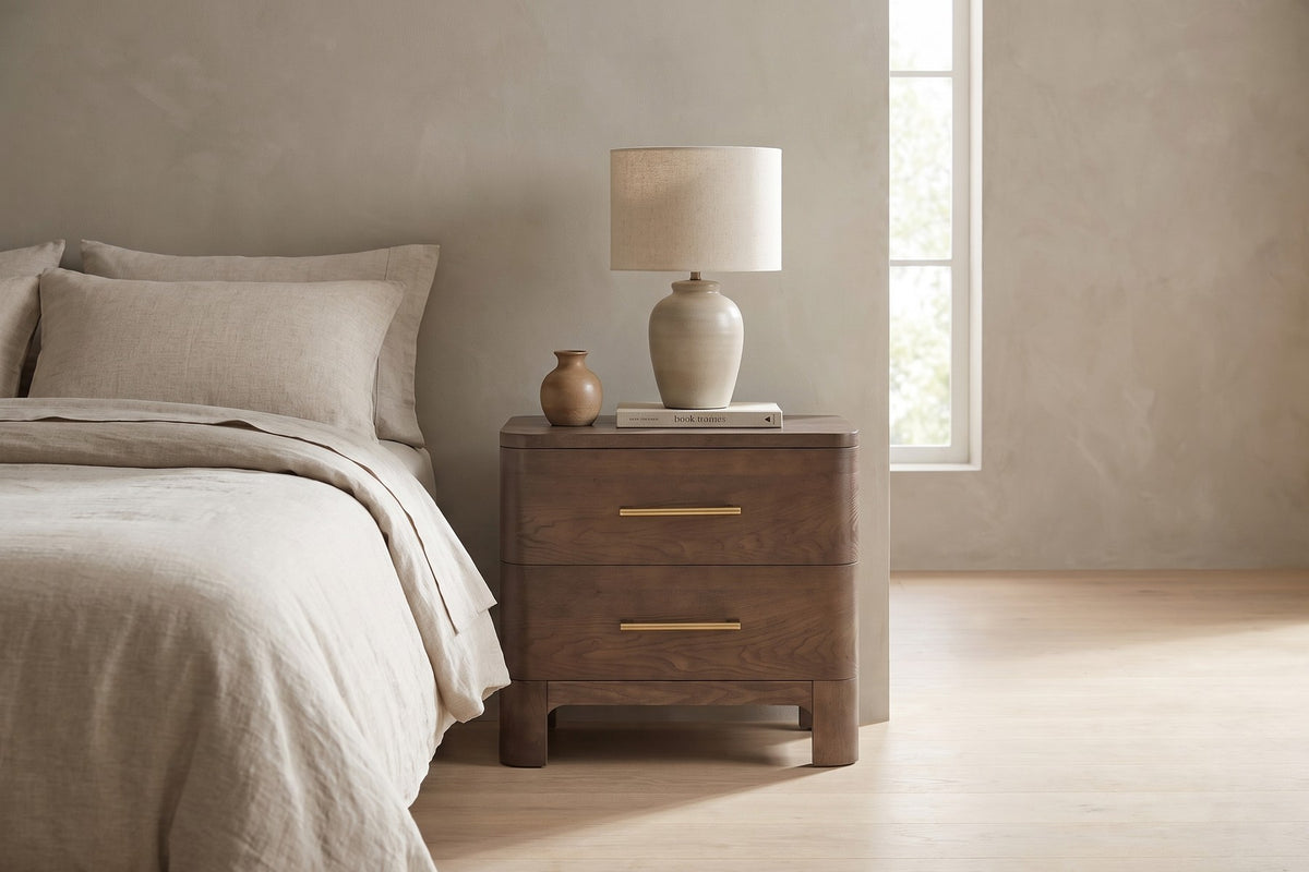 Daylight on the grain. A lamp, a book and a small vase on top, two drawers below.
