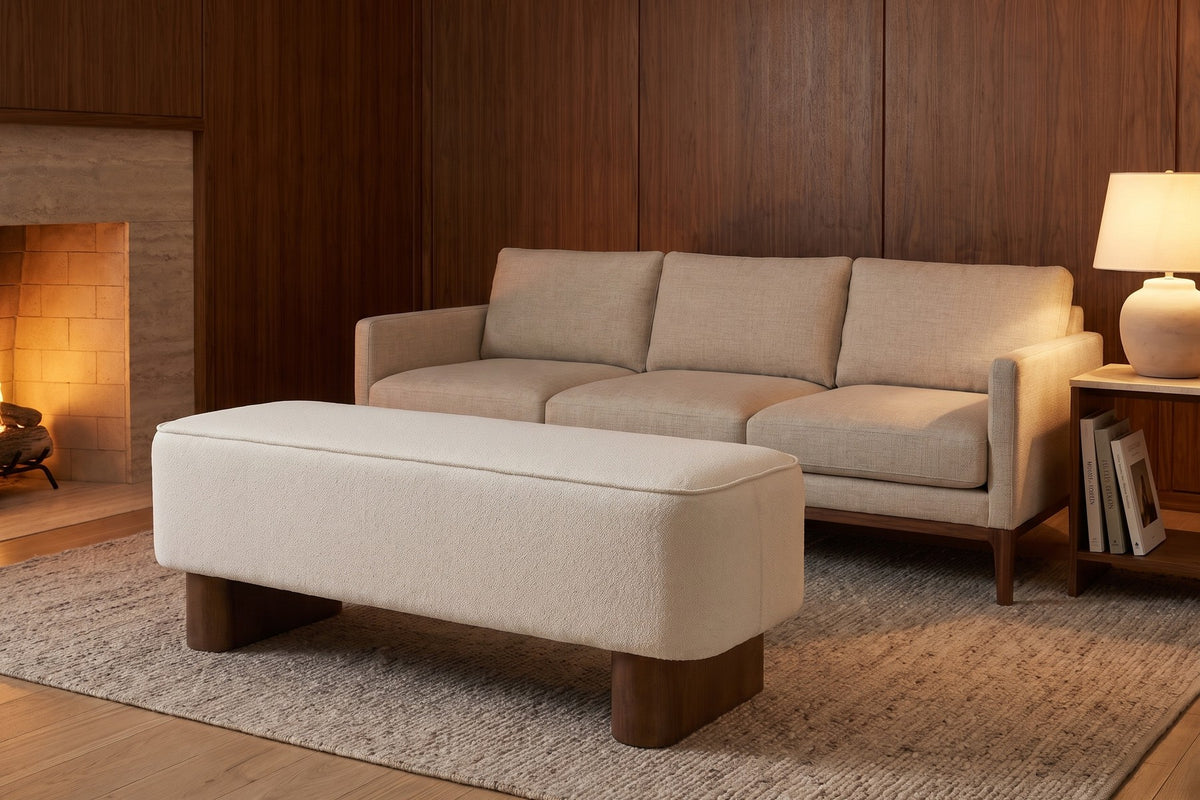 Montsouris 56.5" cream upholstered ottoman bench on rounded rubberwood legs in a warm walnut-panelled living room in the evening
