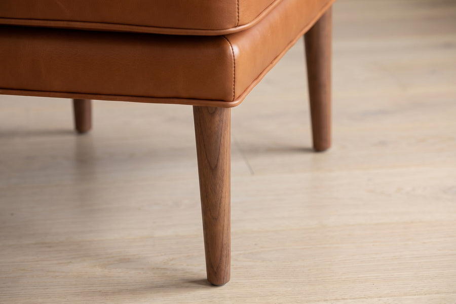 Nyons Cognac Leather Armchair and Ottoman: close-up of a slim tapered solid walnut leg under the front of the ottoman, on a pale oak floor