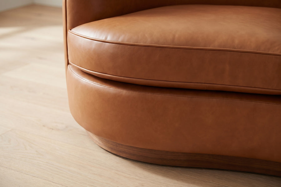 Pornic Cognac Curved Leather Sofa: close-up of the curved front edge of a welted seat cushion and the recessed curved walnut-finish plinth below it, on a pale oak floor