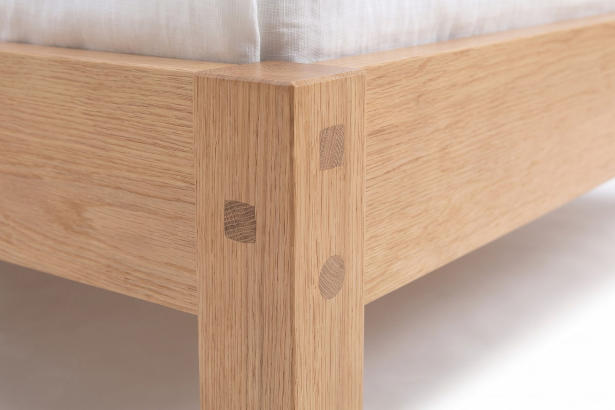 Sancerre Natural Oak Platform Bed: close-up of a foot corner where the oak rail meets the square leg, showing the square end grain of the through-tenon and the round wooden peg