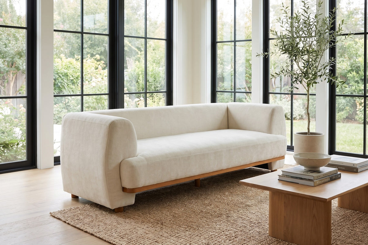 Solange 79" beige chenille sofa with rounded arms and wood base trim in a bright sunroom-style living room