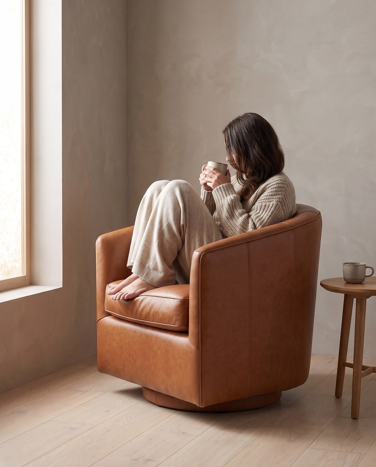 A person curled up with a mug in the Solène cognac leather swivel chair (concept image)