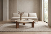 Suzanne 40" round travertine coffee table with dark oak-veneer legs in a calm, daylit living room in front of a sofa