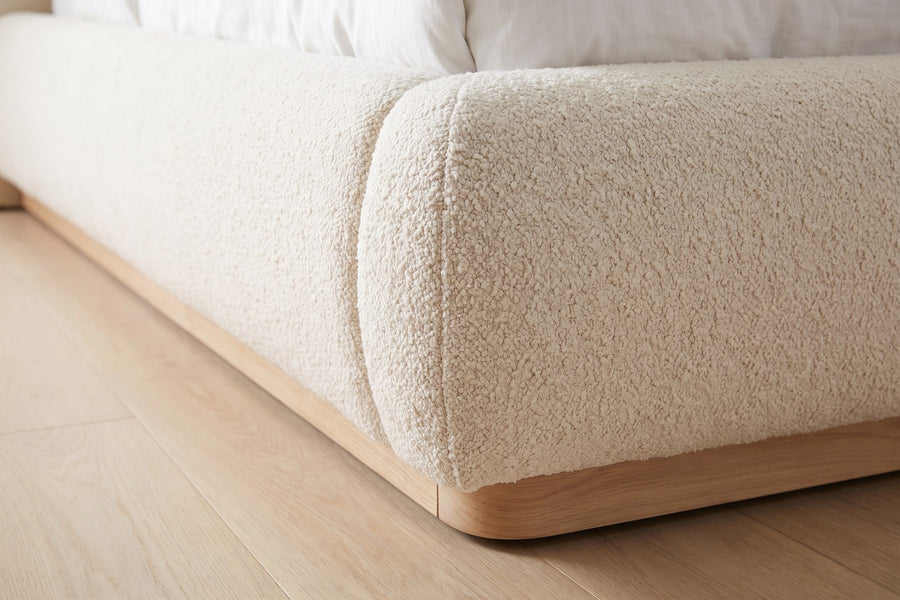 Valloire Ivory Bouclé Bed: close-up of the padded footboard's rounded corner above the solid natural oak plinth, on a pale oak floor
