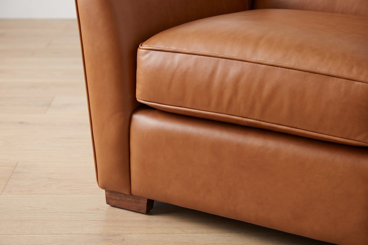 Vence Cognac Leather Lounge Chair: close-up of the welted front edge of the seat cushion and a small walnut-finish block foot below it, on a pale oak floor